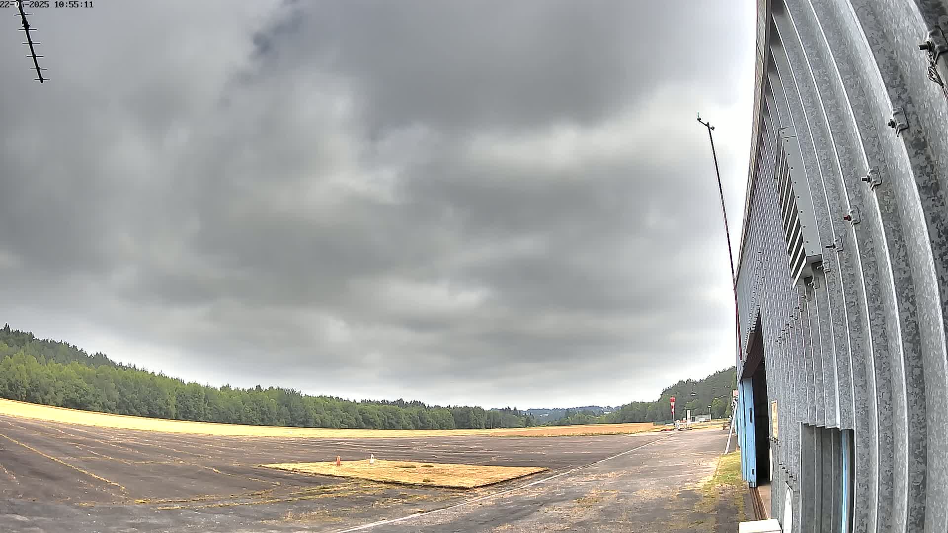 A wide, overcast sky hangs over a flat, paved area bordered by trees, next to a corrugated metal building.