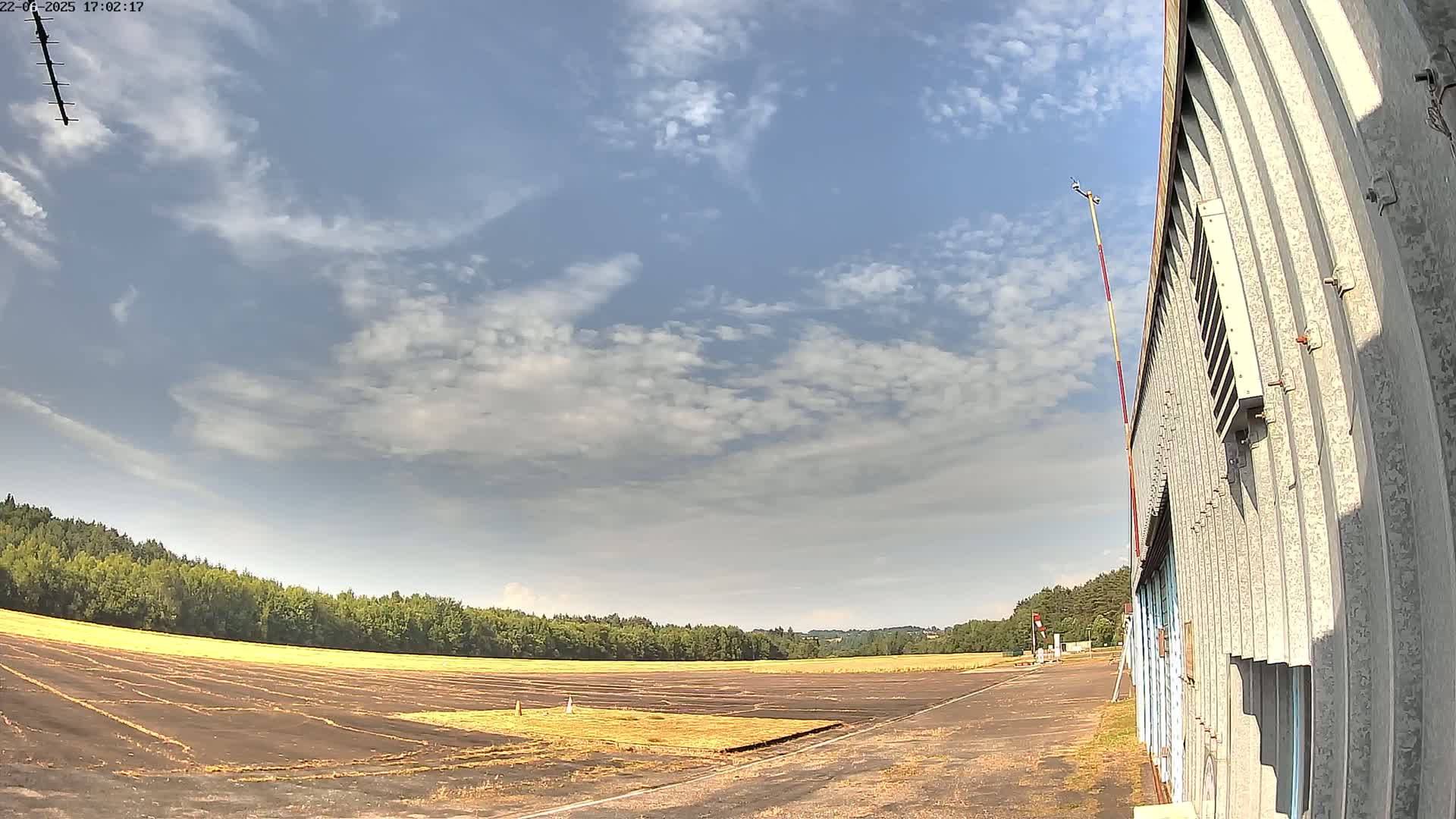 A mostly sunny sky with scattered clouds overlooks a flat, dark brown expanse of land next to a corrugated metal building.