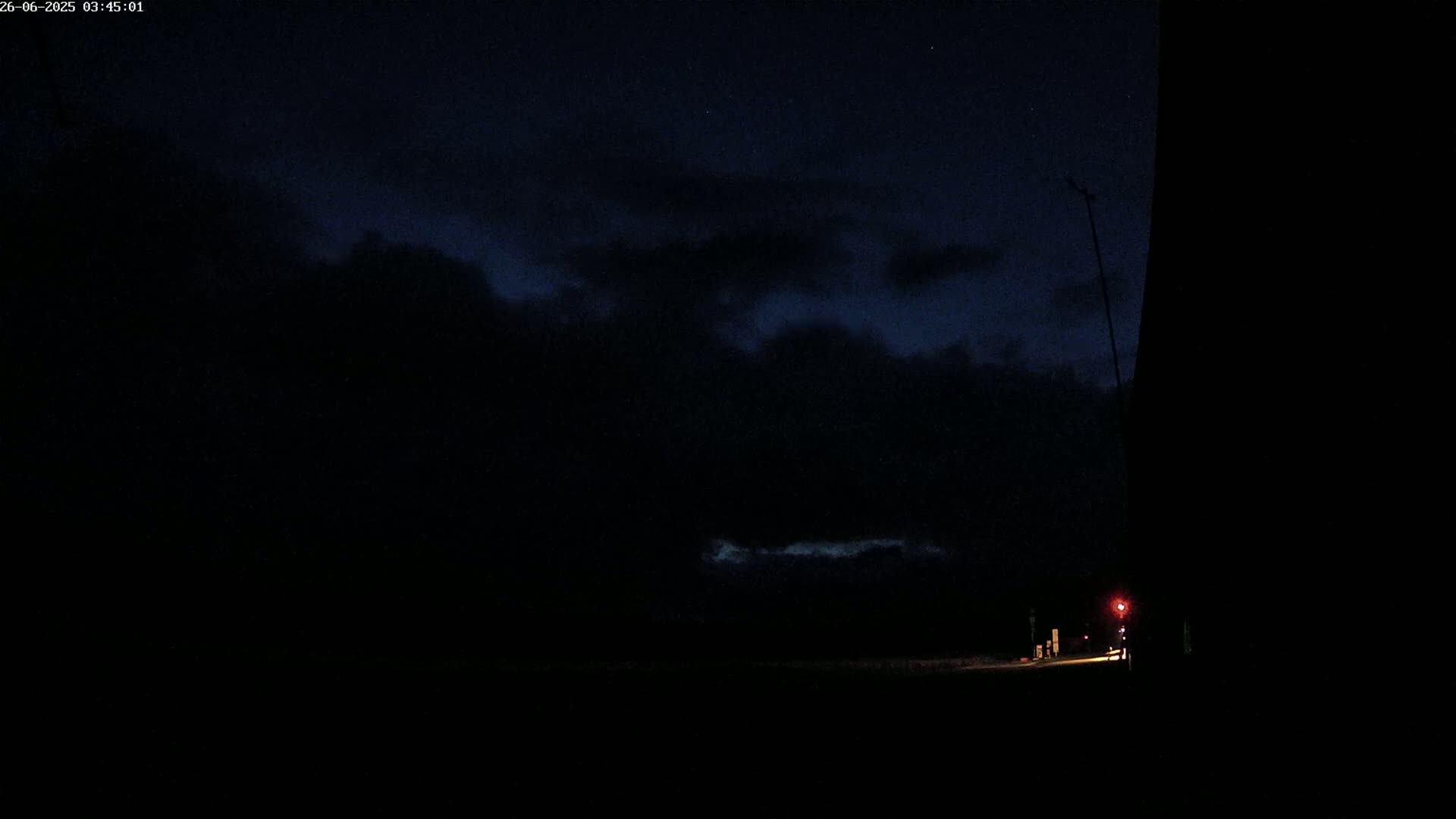 A dark, nighttime scene shows a road with a faint light source in the distance under a cloudy sky.