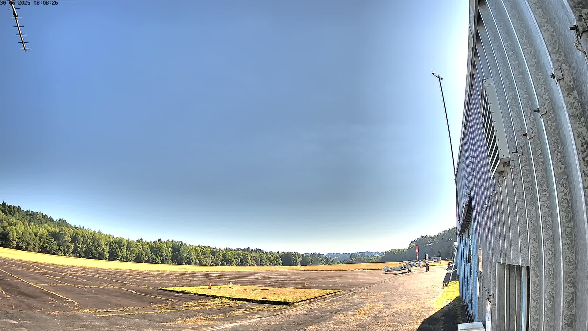 A small airplane sits on a tarmac next to a corrugated metal building under a clear blue sky.
