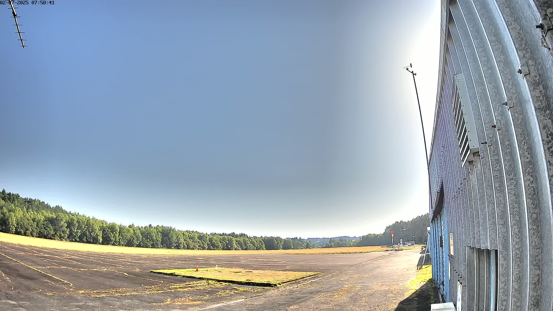 A flat, dark-colored paved area, possibly an airfield, is bordered by a line of trees under a clear, sunny sky, with a corrugated metal building visible to the right.