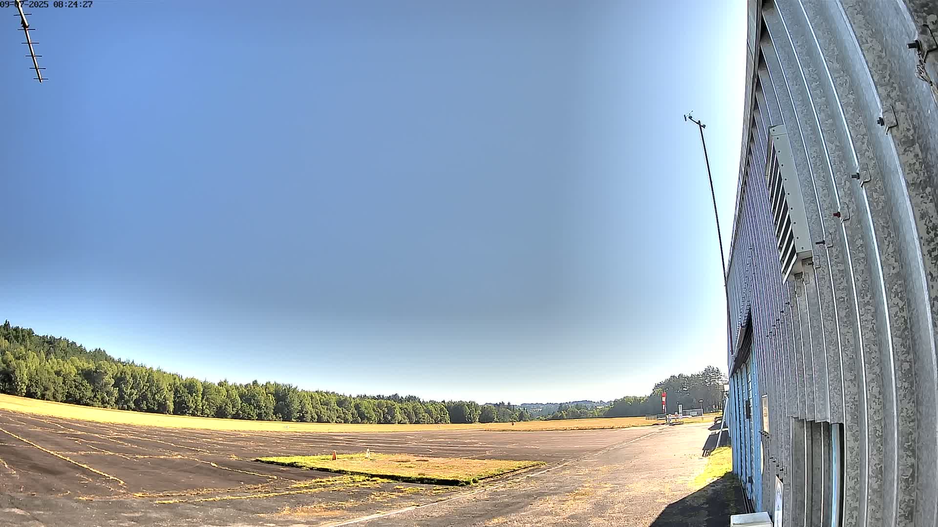 A large, empty tarmac area stretches out before a corrugated metal building under a clear, sunny sky, bordered by a line of green trees.