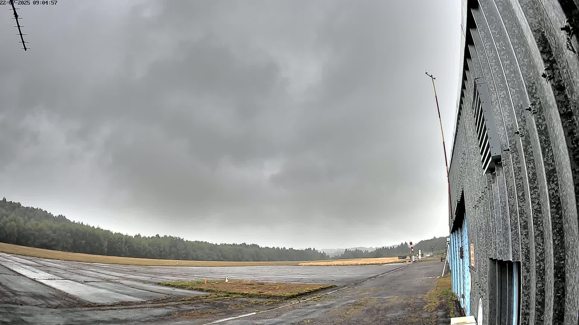 A mostly empty airfield under an overcast sky is seen next to a corrugated metal building.
