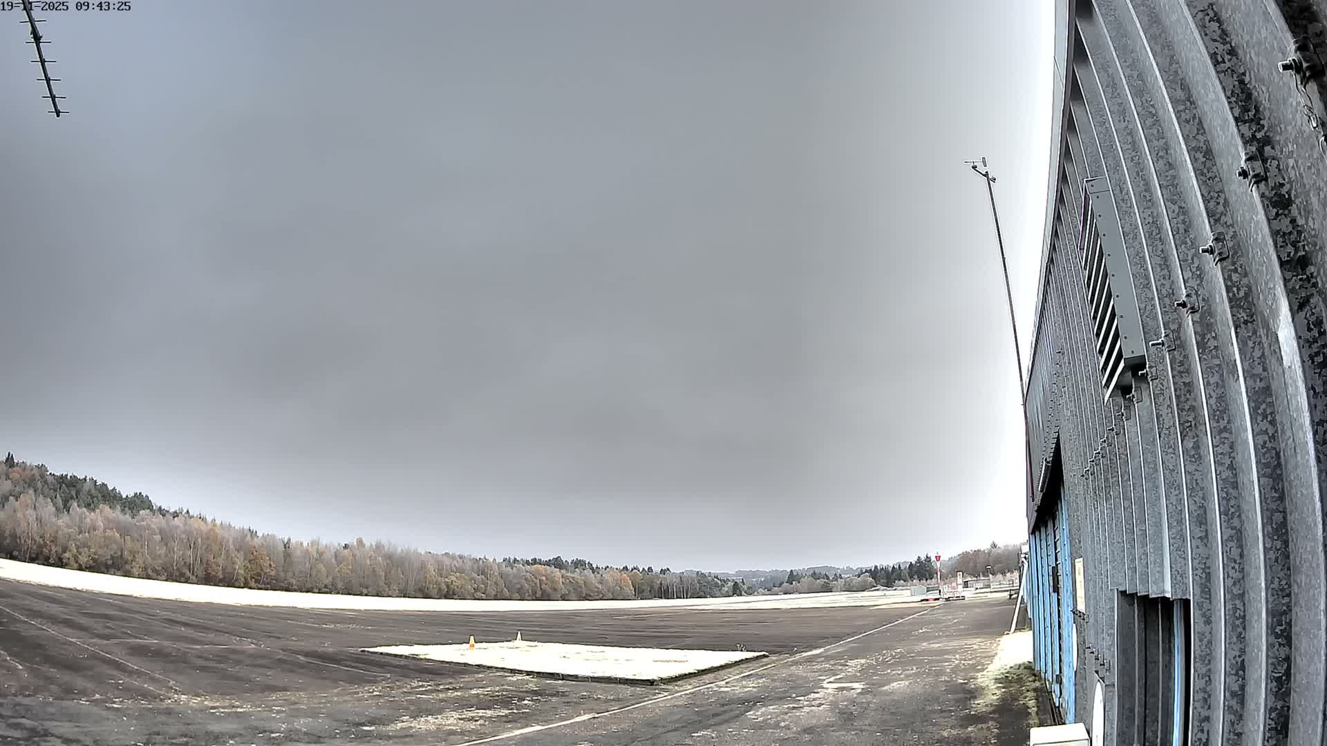 A wide outdoor shot captures a frost-dusted airfield or open paved area bordered by bare trees and an industrial-style building under a uniformly gray, overcast sky.
