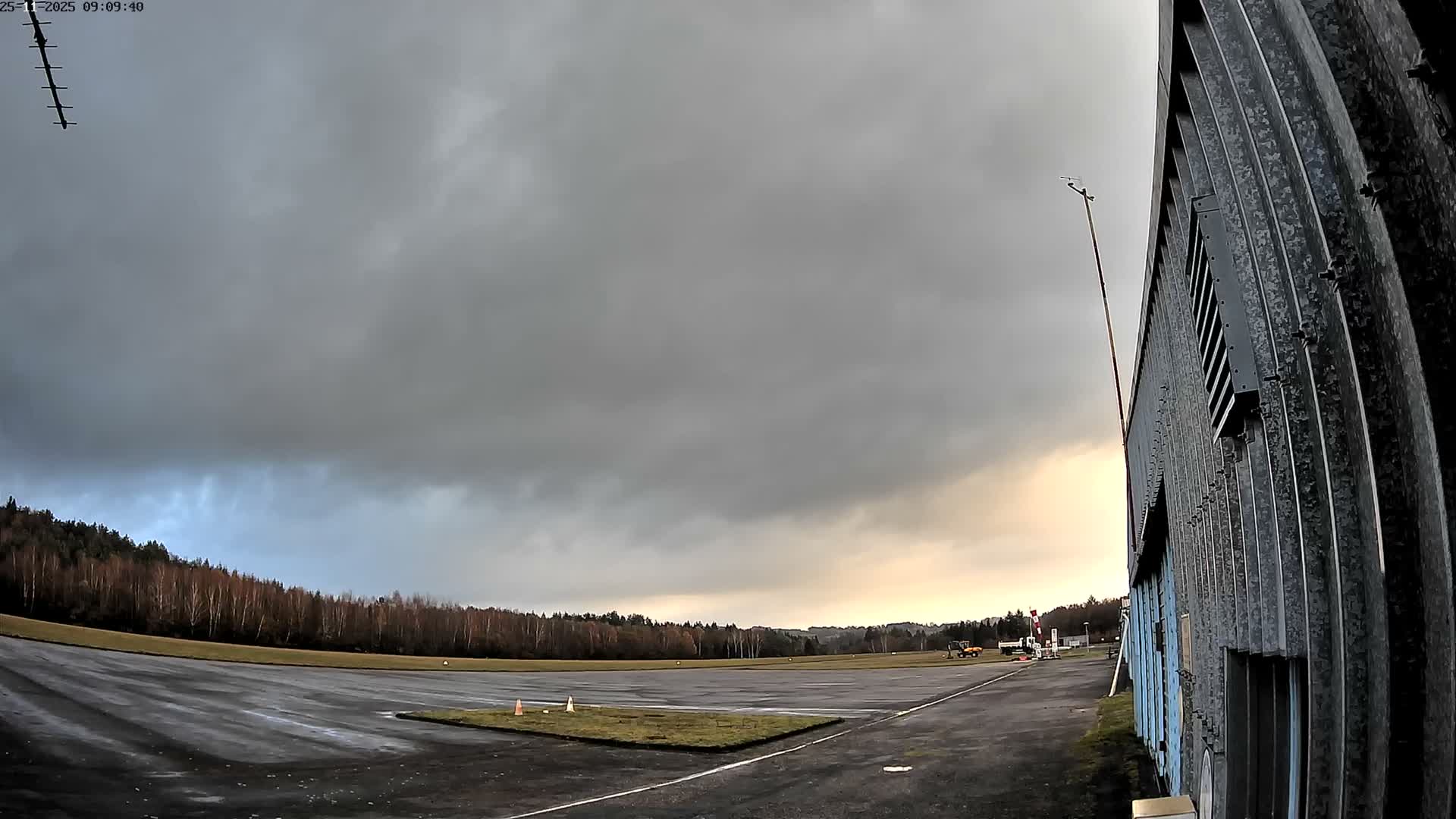 A wide-angle outdoor view captures a wet airfield tarmac bordered by bare winter trees and a corrugated metal building under a heavily overcast and gloomy sky with dark grey clouds.