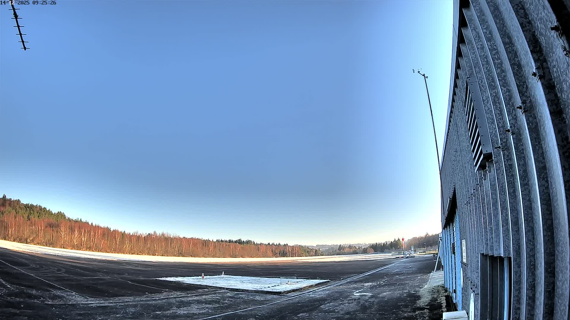 A wide-angle outdoor view captures a wet airfield tarmac bordered by bare winter trees and a corrugated metal building under a heavily overcast and gloomy sky with dark grey clouds.