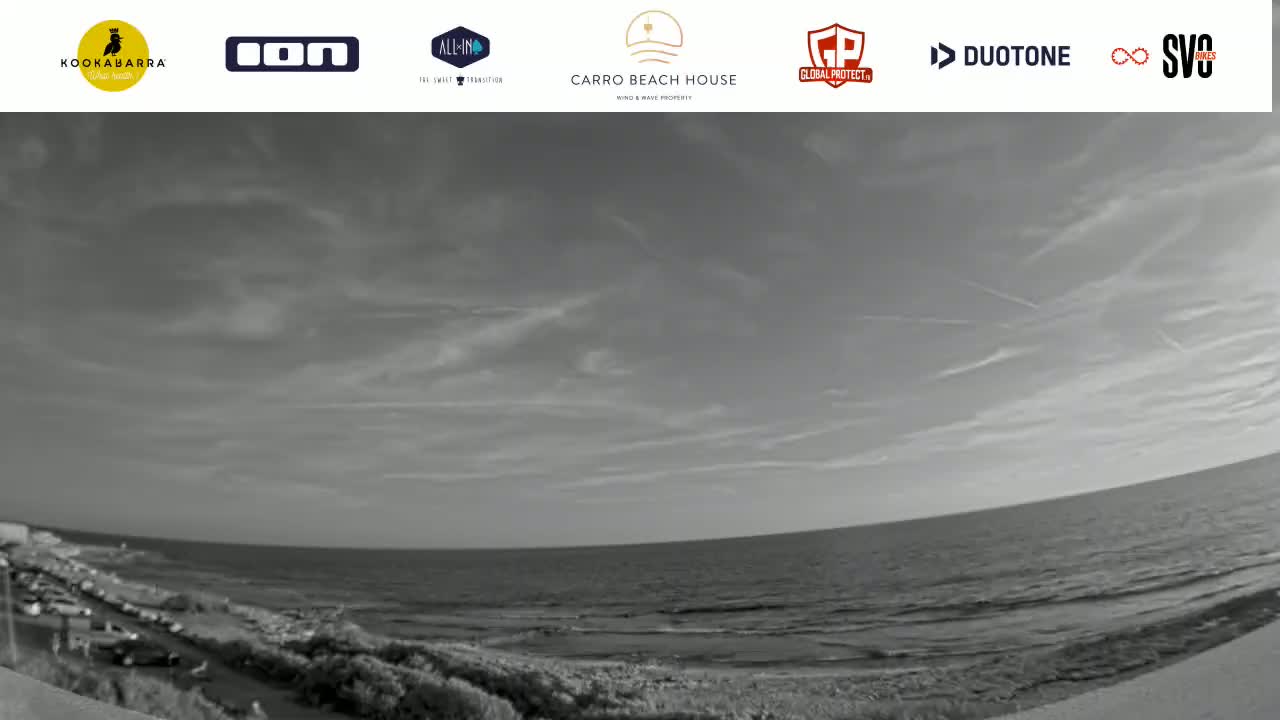 A grayscale, wide-angle view shows a mostly calm ocean meeting a sandy beach, under a partly cloudy sky with some contrails.