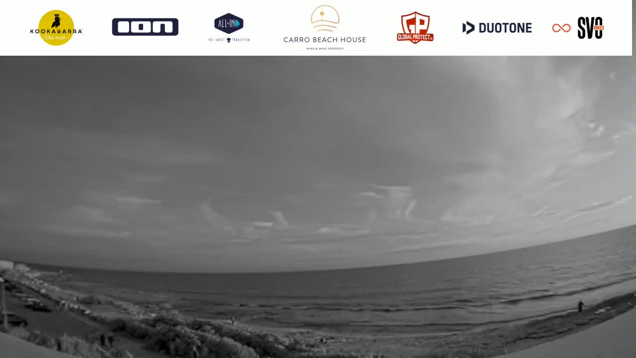 A grayscale wide shot shows a mostly cloudy sky over a calm ocean and beach.