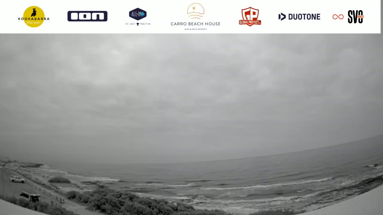 A gray, overcast sky hangs over a calm ocean meeting a sandy shore with sparse vegetation and a few vehicles visible.