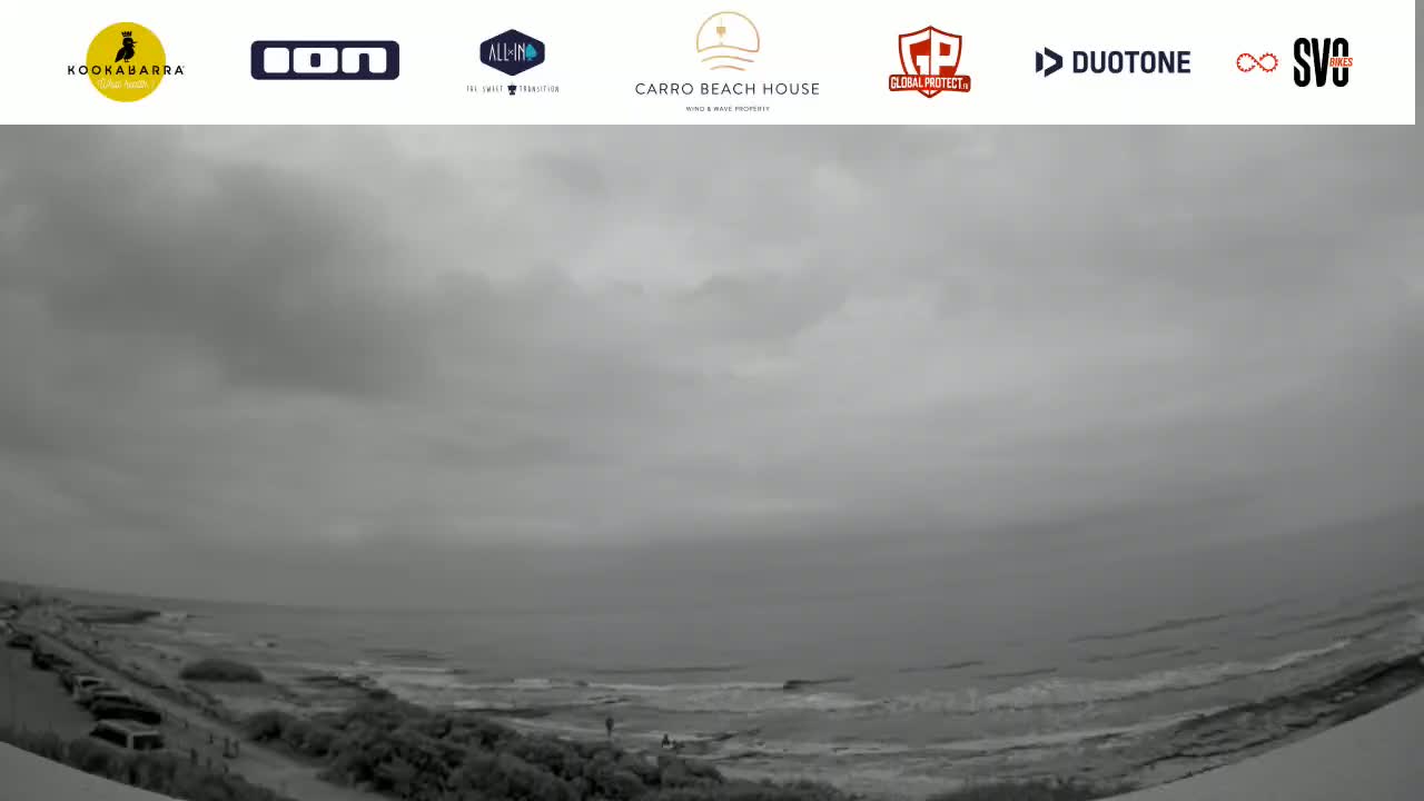 A gray, overcast sky hangs over a beach with calm waves and a few people visible in the distance.