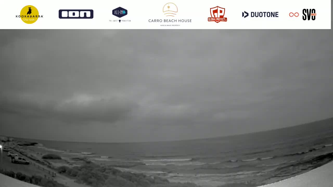 A grayscale image shows a cloudy overcast sky over a calm ocean with small waves lapping a rocky shoreline, where a few cars are parked.