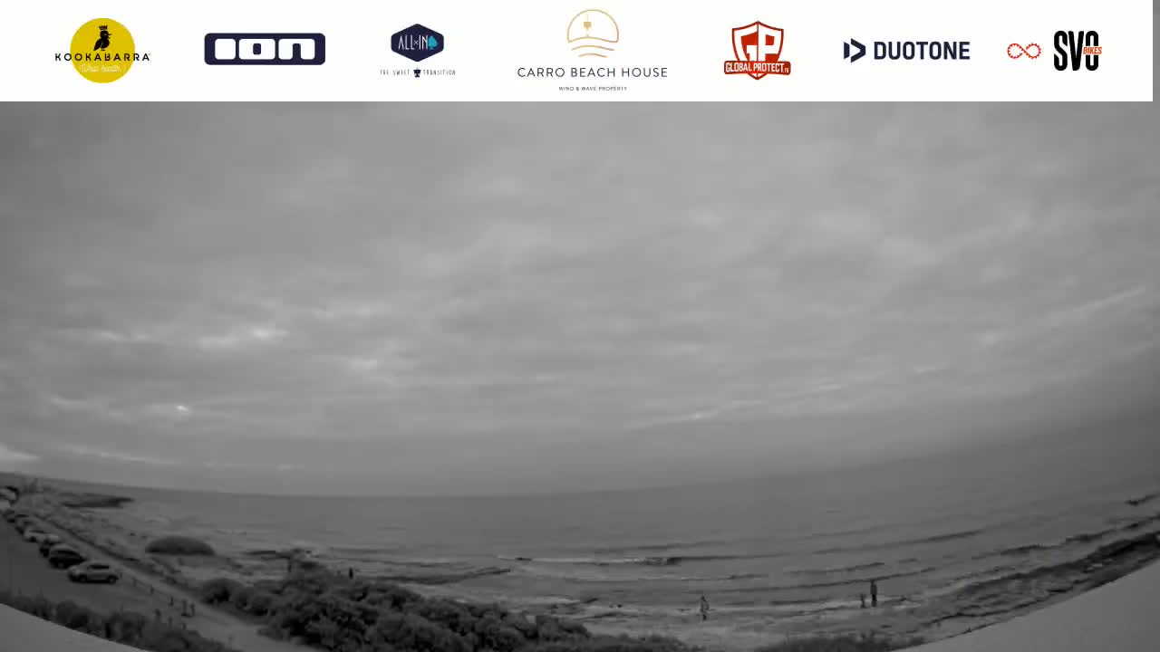An overcast sky hangs over a calm ocean beach scene, with a few people visible near the shoreline.