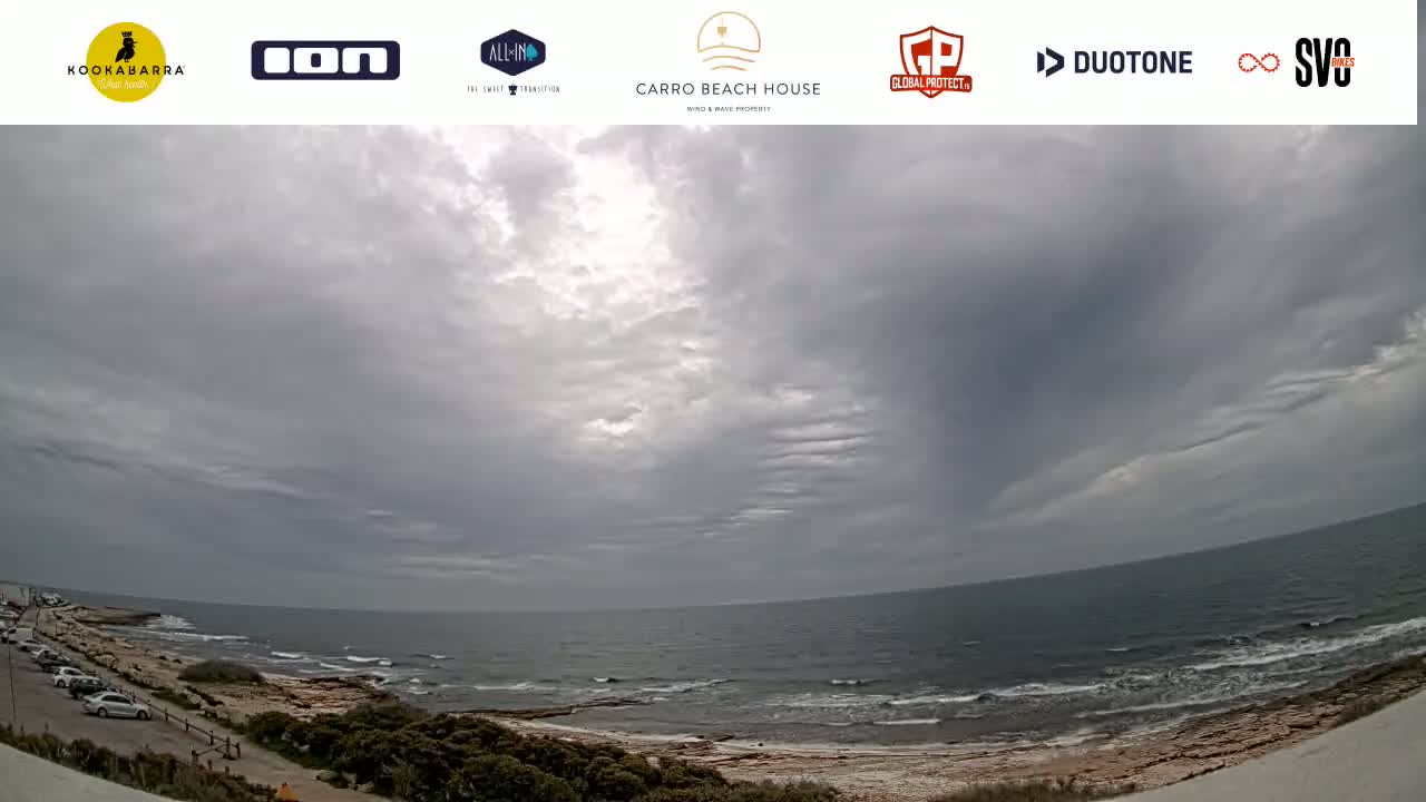 A coastal scene under a mostly cloudy sky shows a rocky shoreline and ocean with small waves.