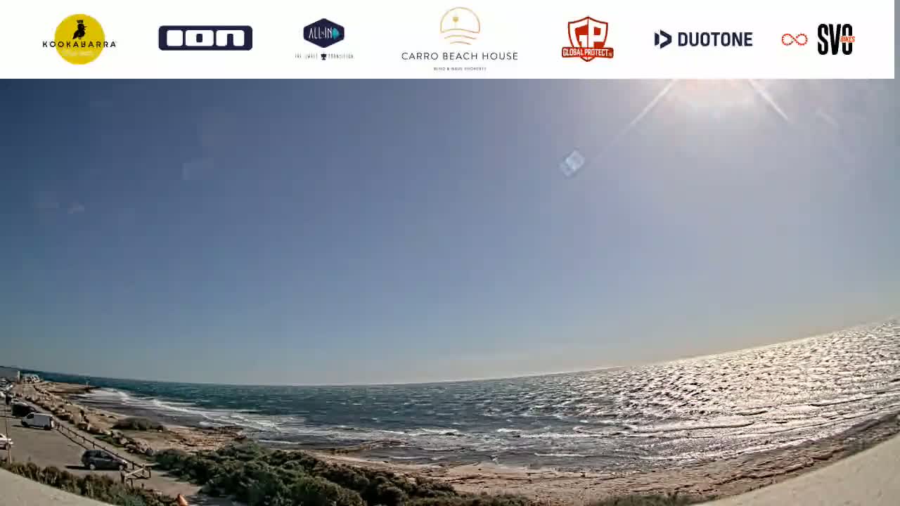 A sunny day at the beach shows a rocky shoreline, a wavy ocean, and a few cars parked nearby.