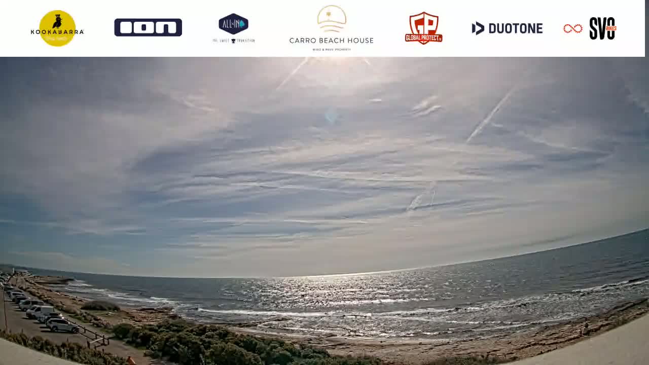 A mostly sunny day reveals a wide, gently wavy ocean meeting a sandy shore with parked cars and sparse vegetation visible in the foreground.