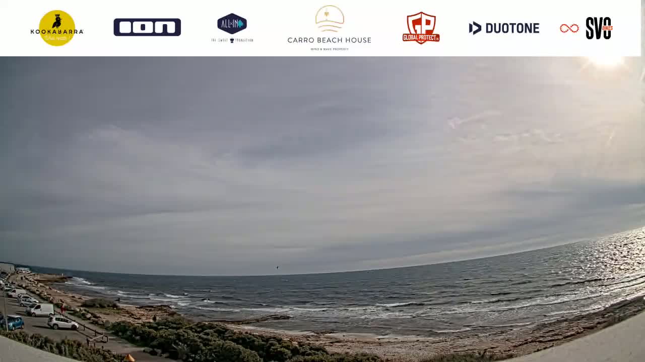 A mostly cloudy day shows a wide, sandy beach with a calm ocean and a few cars parked nearby.