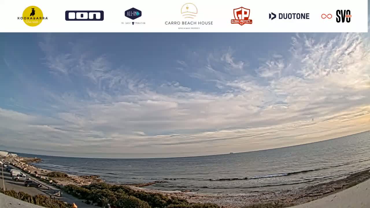 A wide shot of a calm ocean under a mostly clear sky with thin, wispy clouds shows a rocky shoreline with a parking lot and road.
