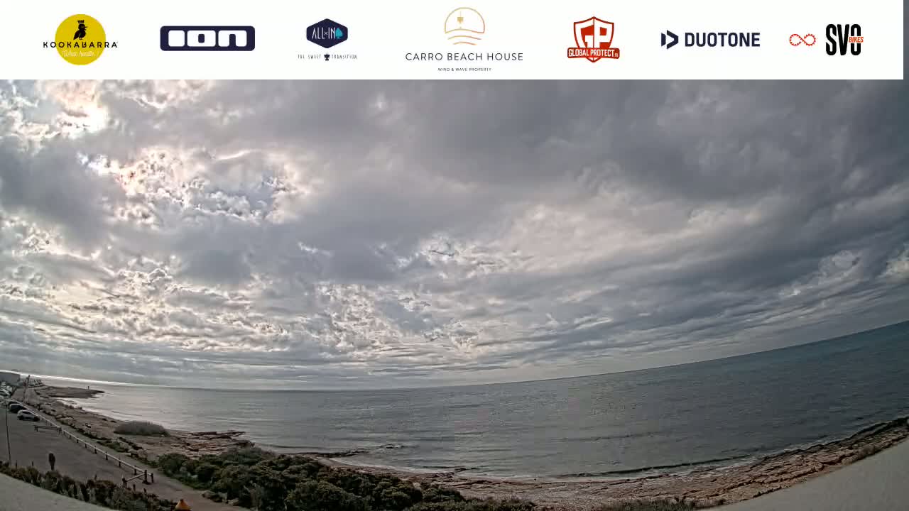 A mostly cloudy sky hangs over a calm ocean meeting a rocky shoreline with sparse vegetation.