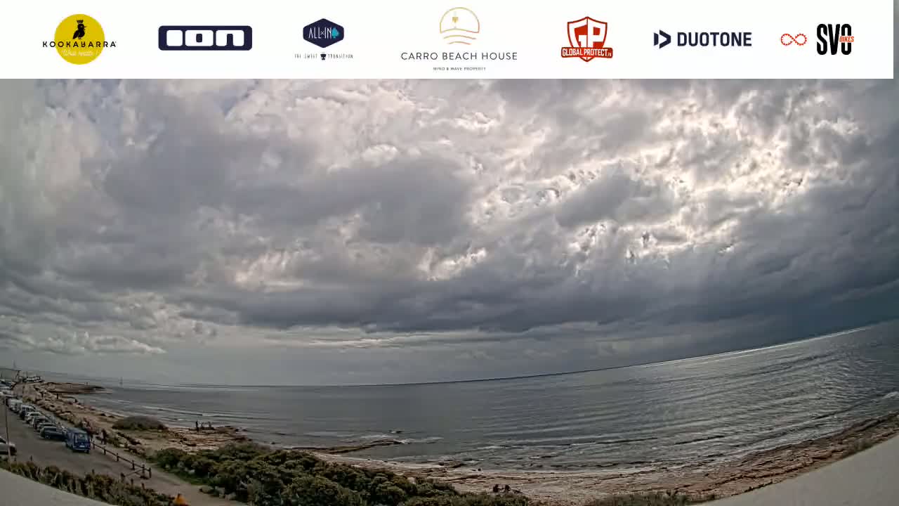 A wide, overcast sky hangs over a calm ocean and a rocky shoreline with a few people and parked cars visible.
