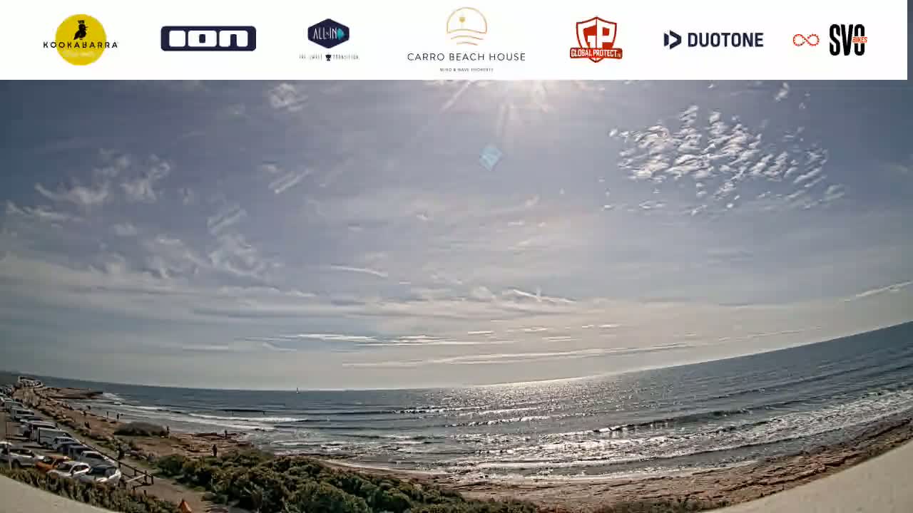 A sunny day with partly cloudy skies overlooks a beach with ocean waves and parked cars.