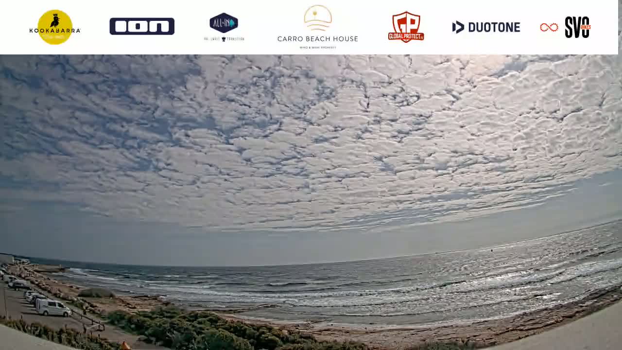 A mostly sunny day with scattered clouds shows a wide expanse of ocean with small waves lapping a sandy shore, backed by a coastal road with parked cars and vegetation.