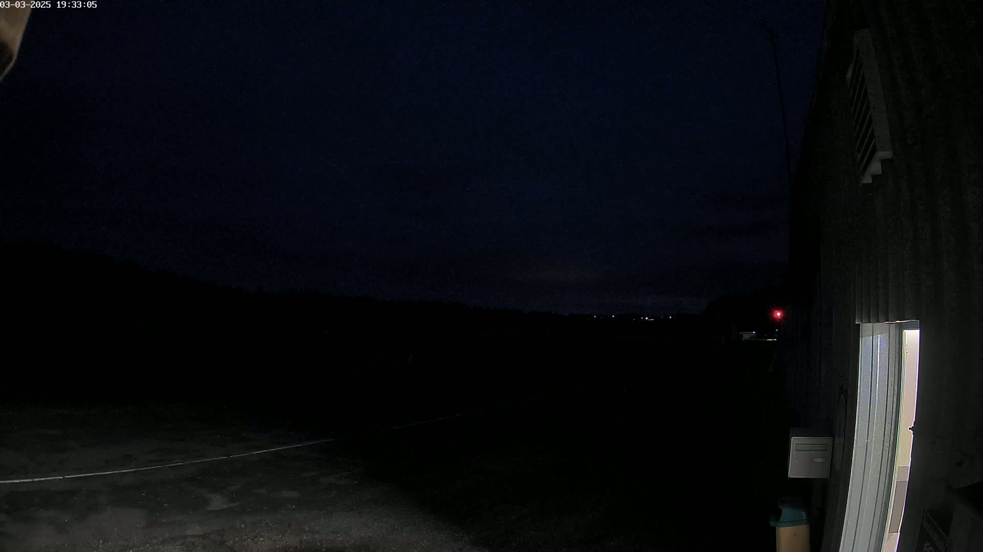 A dark, nighttime scene shows a paved area next to a building with a slightly open door, under a mostly clear, dark sky.