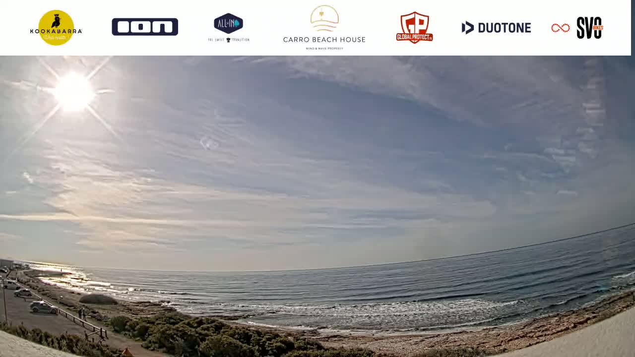 A sunny day with a mostly clear sky shows a wide view of a calm ocean meeting a sandy shore with some vegetation and a few people visible.