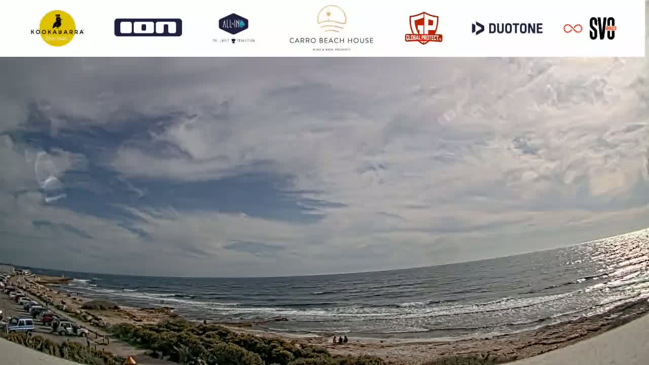A partly cloudy sky overlooks a beach with a calm ocean and a few people visible near the shoreline, with cars parked in a lot nearby.