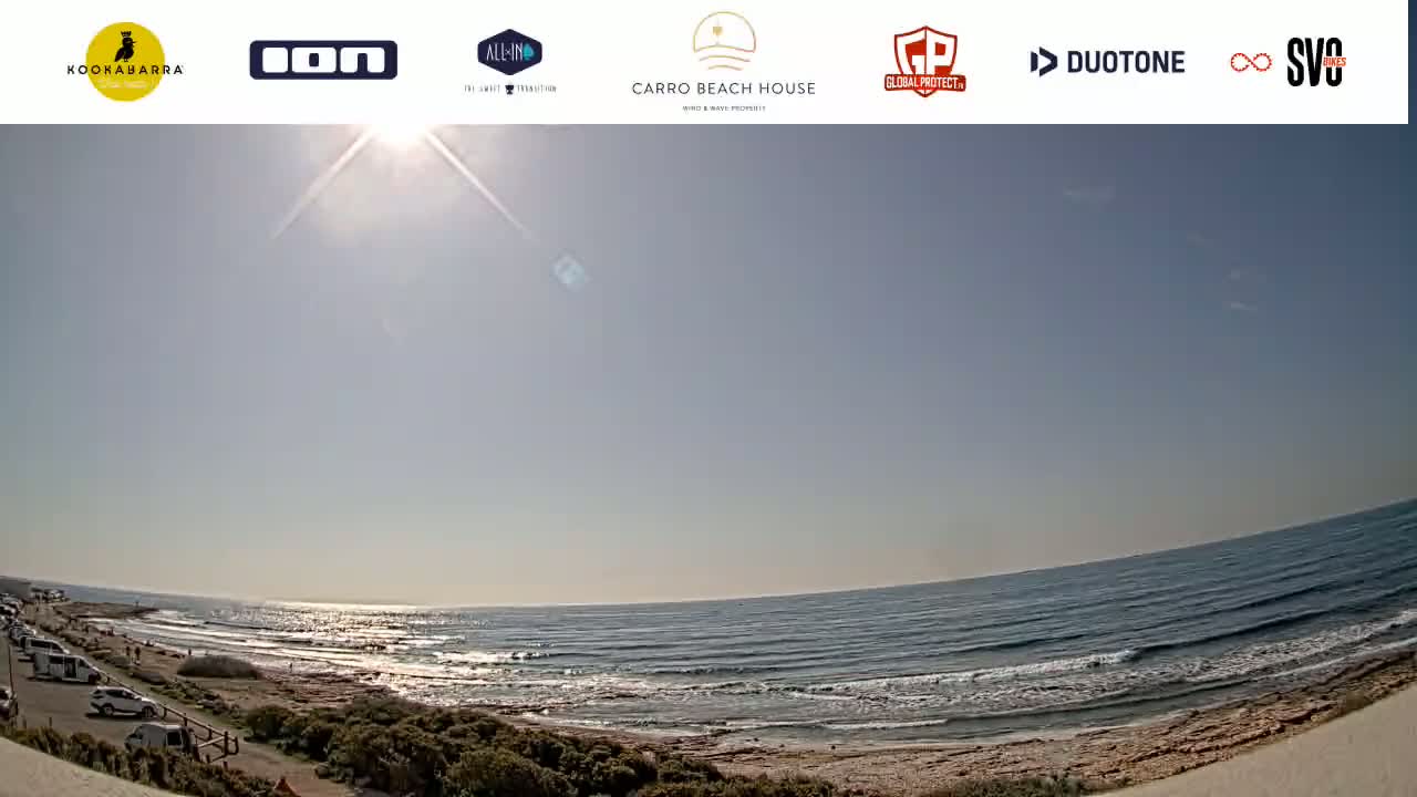 A sunny, mostly clear day at a beach with a parking lot and some small waves rolling onto the shore.