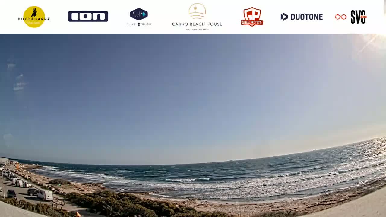 A mostly sunny day at a beach with moderate waves, showing a parking lot with several cars and a shoreline with some vegetation.