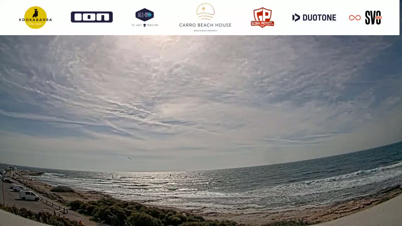 A mostly sunny day with scattered clouds over a beach with calm waves and several cars parked along the shore.