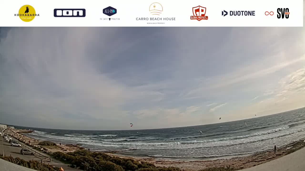 Several kitesurfers are visible on a windy day at an ocean beach with a mostly cloudy sky.