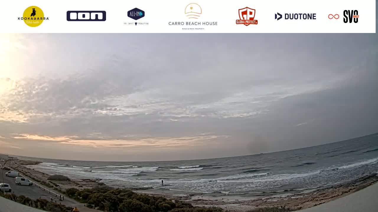 A wide shot of an overcast beach scene at sunset shows a calm ocean with small waves breaking on a rocky shore, with cars parked along a road in the distance.