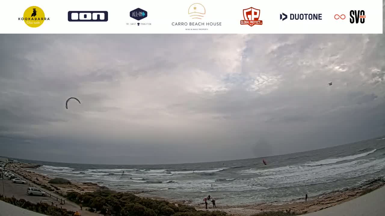 A kiteboarder and windsurfers are visible on a somewhat overcast day at a beach with a rocky shoreline and small waves.