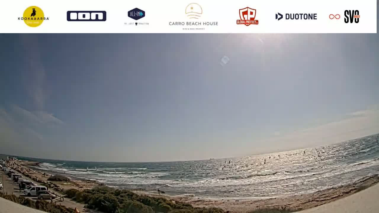 A sunny day at a beach shows a mostly calm ocean with some small waves and several people windsurfing in the distance, with a parking lot and some vegetation visible along the shore.