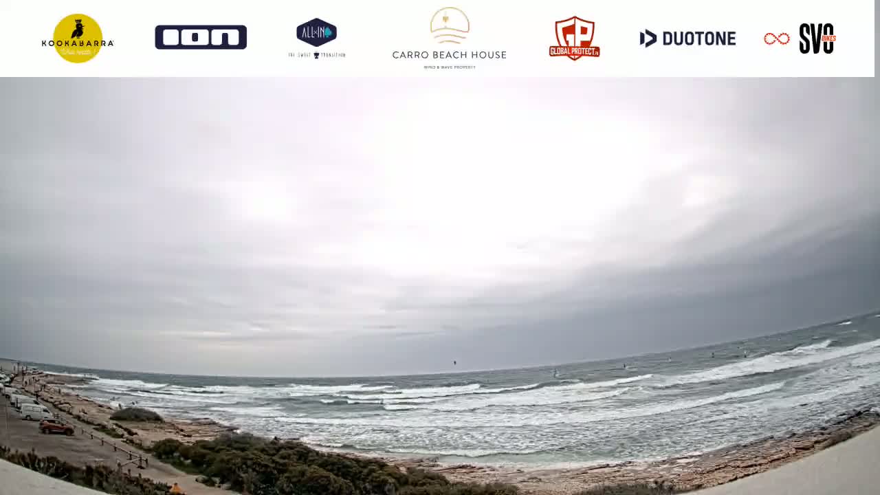 A wide shot shows a rocky beach with a few people and parked cars under an overcast sky, overlooking a choppy ocean with several windsurfers.