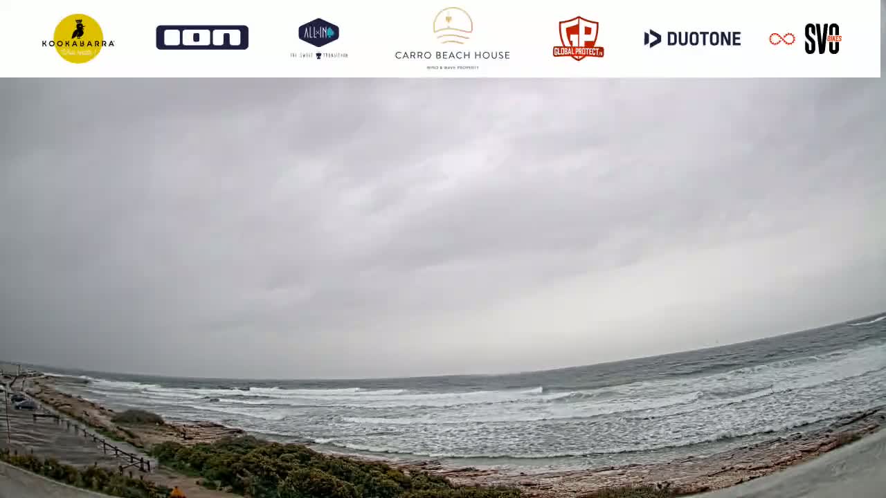 A wide, overcast view shows a rocky coastline with small waves lapping the shore under a gray sky.