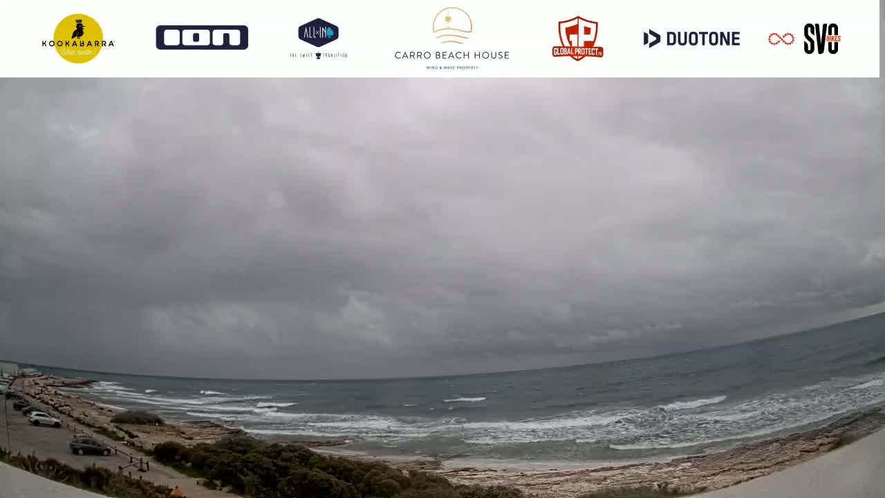 A wide shot shows a partly cloudy, overcast sky over a beach with a rocky shoreline and small waves rolling into shore.