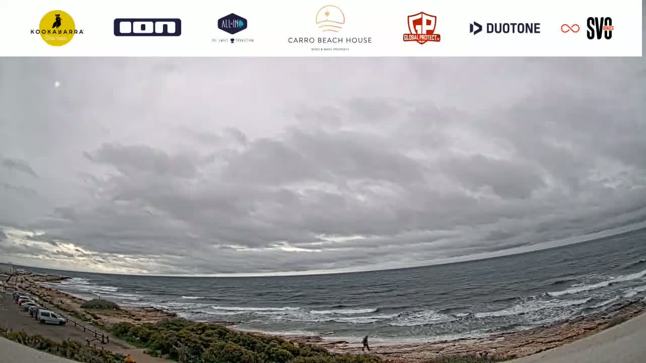 A person walks along a rocky shoreline next to a dark, choppy ocean under a mostly cloudy sky.