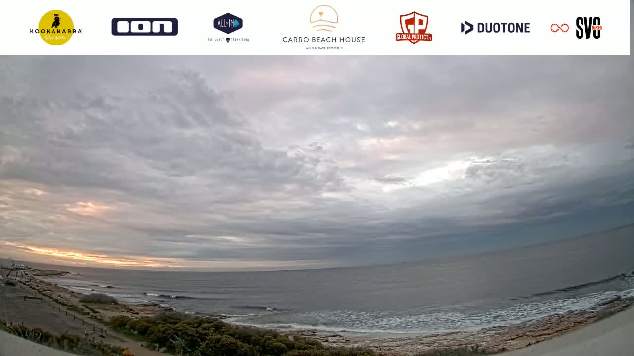 A wide shot shows a mostly cloudy sky over a calm ocean with small waves breaking on a rocky shore at sunset.