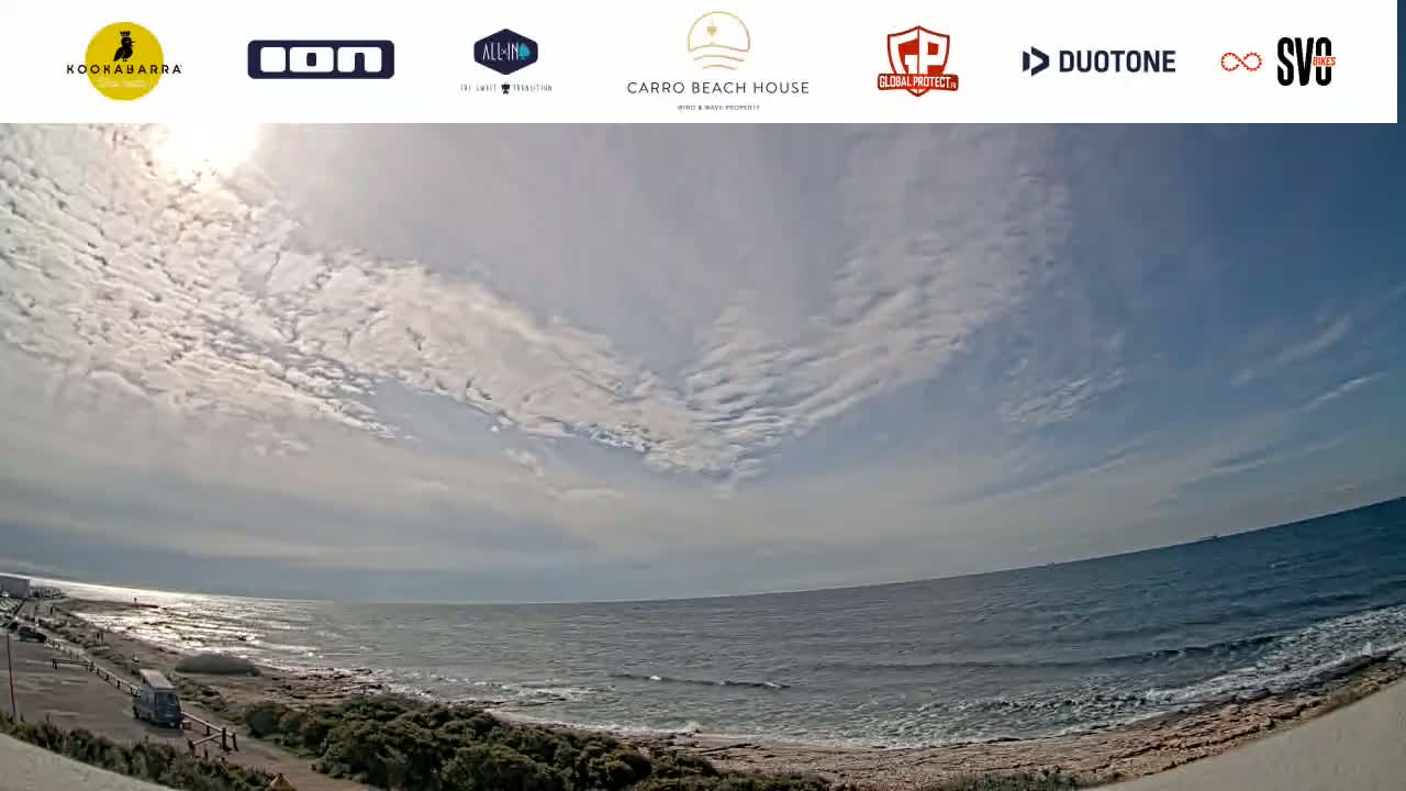 A wide shot of a beach with a partly cloudy sky shows a calm ocean with small waves lapping the shore.