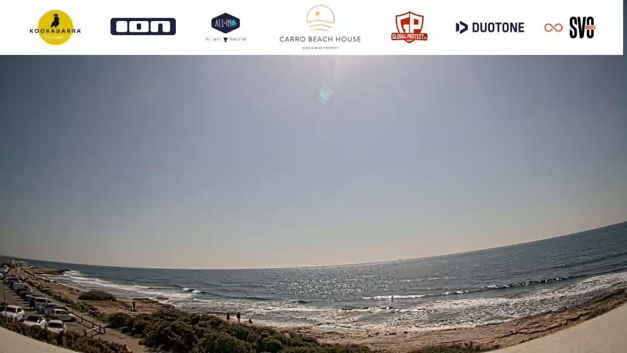 A sunny day at a beach shows a parked lot, a sandy shore, and a calm ocean with small waves.