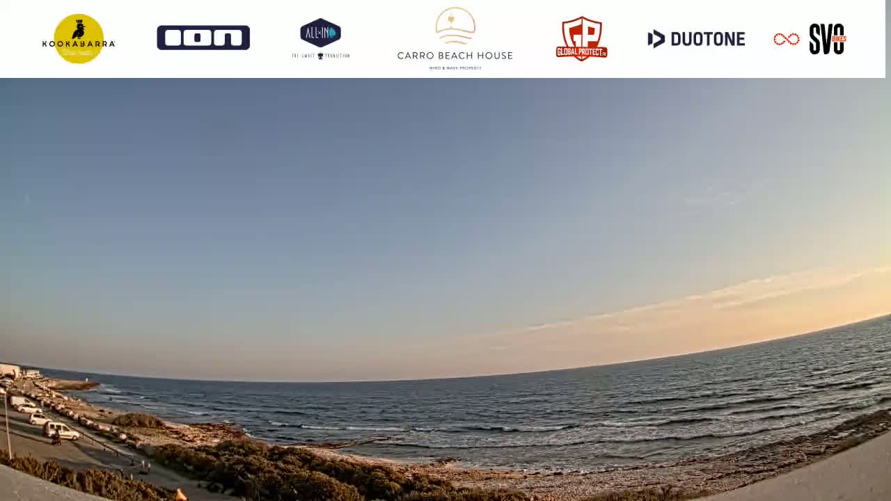 A calm ocean with small waves meets a sandy shore lined with sparse vegetation under a clear, pale-orange sunset sky.