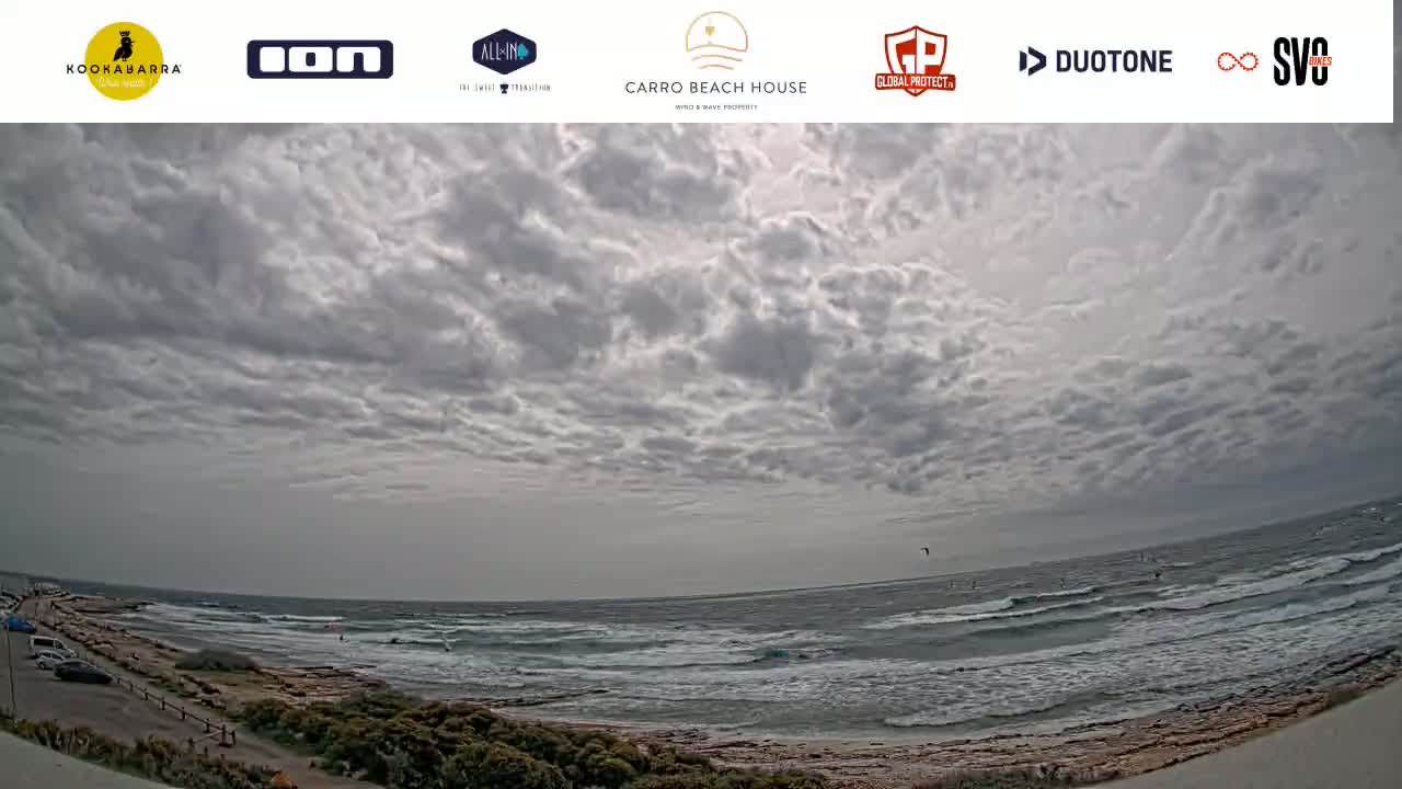 A wide shot shows a partly cloudy, overcast sky above a rocky coastline with a wavy ocean where several kitesurfers are visible in the distance.