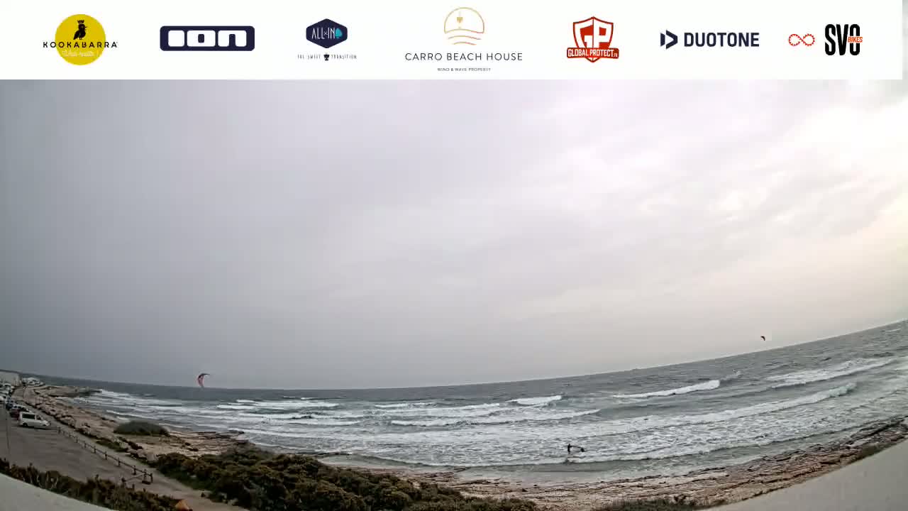 An overcast sky hangs over a wavy ocean, with a few kitesurfers visible in the distance and some people on the shore.