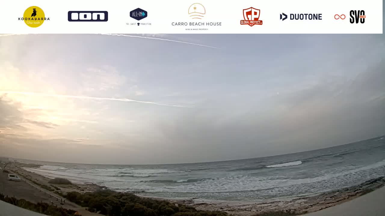 A hazy sunrise illuminates a wide, somewhat choppy ocean meeting a sandy shoreline with sparse vegetation.
