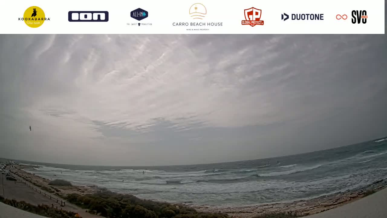 A mostly cloudy day reveals a rocky coastline with a choppy ocean where several people are windsurfing or kitesurfing.