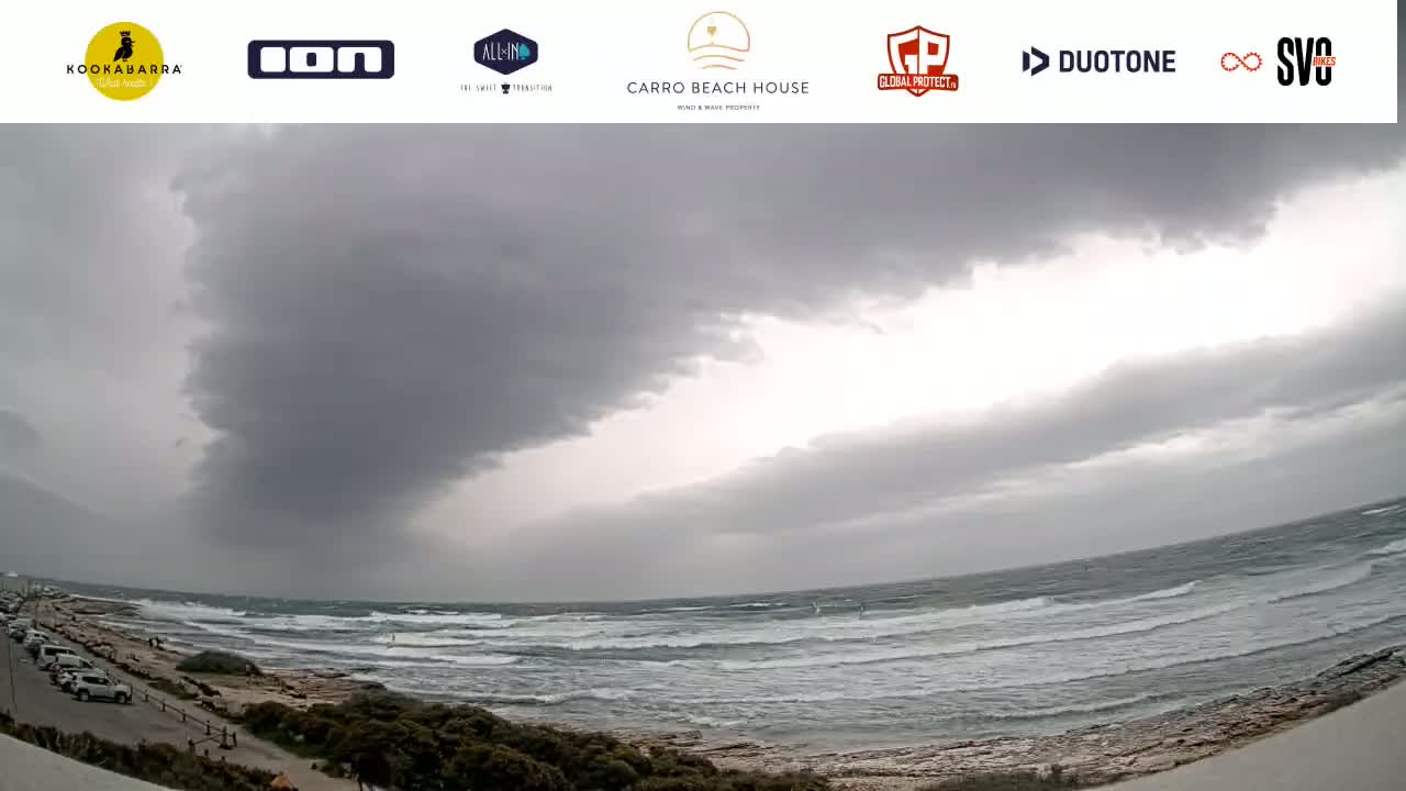A dark, overcast sky hangs over a choppy ocean, with a beach and a parking lot visible in the foreground.