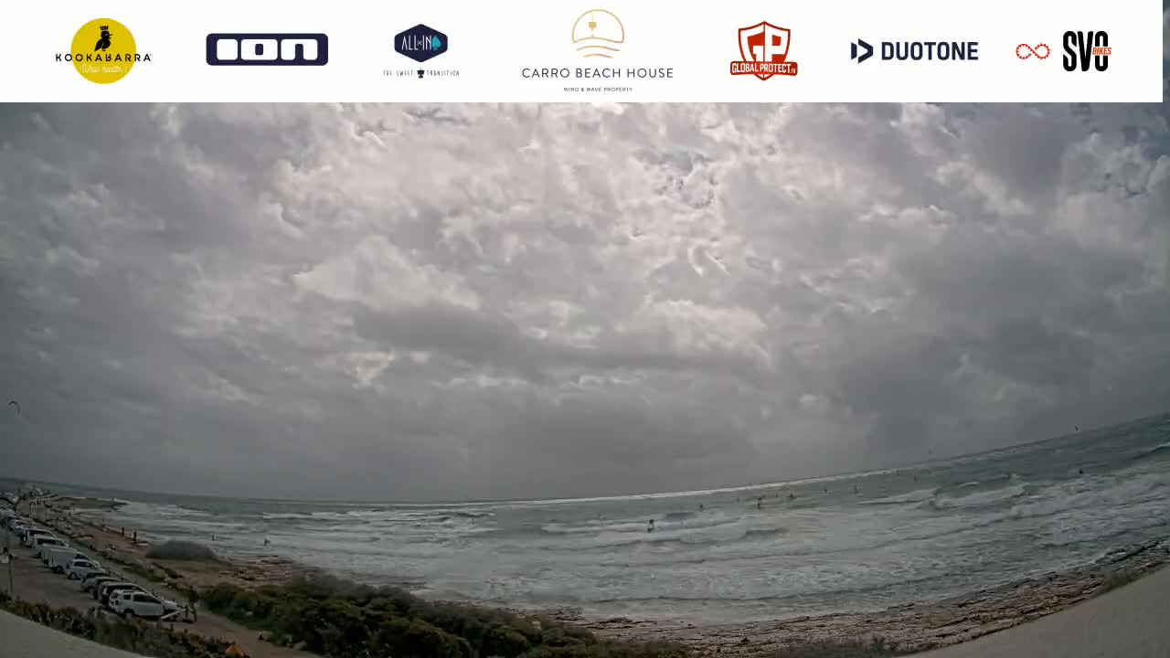 A wide shot of a beach with overcast skies and moderate waves, showing several people in the water.
