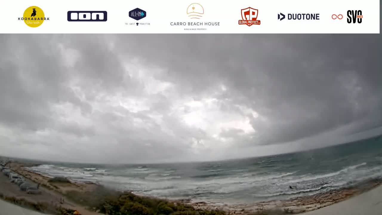 A wide shot shows a stormy, overcast sky over a choppy ocean meeting a sandy shore with some vehicles parked nearby.